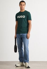 T-shirt verde de algodão com grande texto branco "HUGO", combinado com jeans azul claro e sapatilhas bege; bolsa preta com alça.