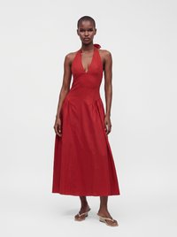 Woman wearing a sleeveless red halter neck midi dress and beige heeled sandals, standing against a white background.