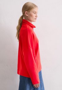 Rode sweatvest met ritssluiting, hoge kraag en losse pasvorm, voorzien van lange mouwen. Gecombineerd met een denim rok.