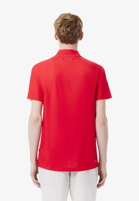 Röd kortärmad polo med ribbad krage, slät textur, med en etikett som har "Lacoste"-logotypen bak. Enfärgad design.