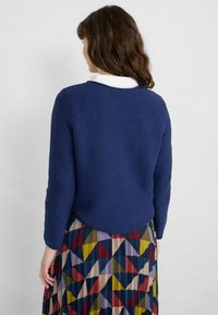 Maglione lavorato a maglia blu navy con scollo rotondo e maniche lunghe, abbinato a una gonna plissettata con motivi geometrici in più colori.