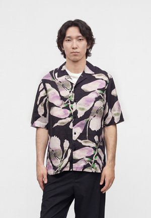 Jeune homme aux cheveux noirs mi-longs portant une chemise noire à manches courtes avec de grands motifs floraux violets et blancs.
