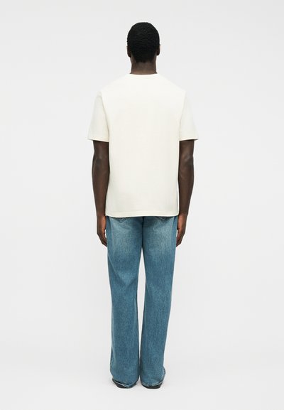 Άνδρας που στέκεται πλάτη φορώντας απλό λευκό κοντομάνικο πουκάμισο και άνετο μπλε τζιν, μπροστά σε απλό λευκό φόντο.