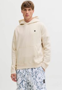 Haut à capuche beige avec une poche kangourou sur le devant et un petit logo noir sur la poitrine. Le tissu semble doux et le design a une coupe décontractée.