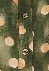Grøn skjorte med knapper og en subtil glans, prydet med store beige polka-prikker og en metallisk knap. Glat stof med en let tekstureret overflade.