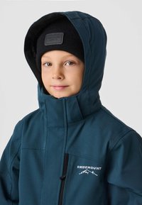 Morska zielona kurtka przeciwdeszczowa z wysokim kołnierzem i kapturem, z czarnym zamkiem oraz kieszenią na piersi z logo "Swedemount". Pod spodem czarna czapka beanie.