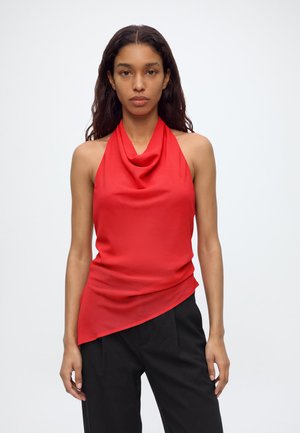 Femme aux longs cheveux foncés portant un haut rouge sans manches à col bénitier et un pantalon noir, debout sur un fond clair uni.