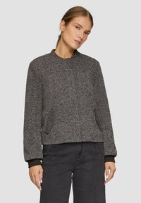 Veste en chevrons grise avec un col rond, des manches longues et des poches latérales. Dispose de poignets et d'un ourlet en tricot noir. Portée sur un jean foncé.