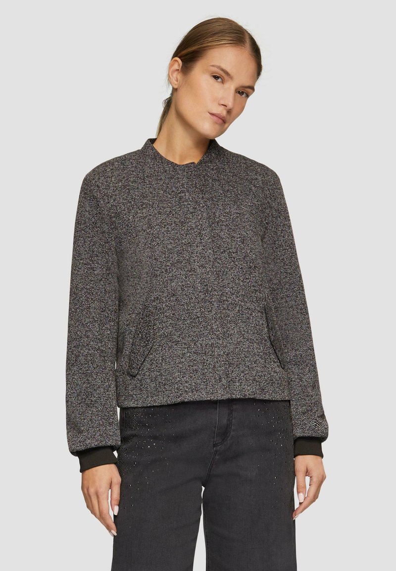 Veste en chevrons grise avec un col rond, des manches longues et des poches latérales. Dispose de poignets et d'un ourlet en tricot noir. Portée sur un jean foncé.