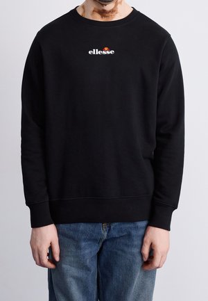 Homme portant un sweat-shirt noir avec un petit logo « ellesse » sur la poitrine et un jean bleu, debout devant un fond clair uni.