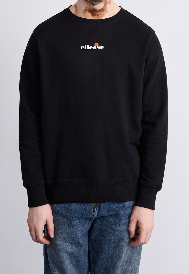 Homme portant un sweat-shirt noir avec un petit logo « ellesse » sur la poitrine et un jean bleu, debout devant un fond clair uni.