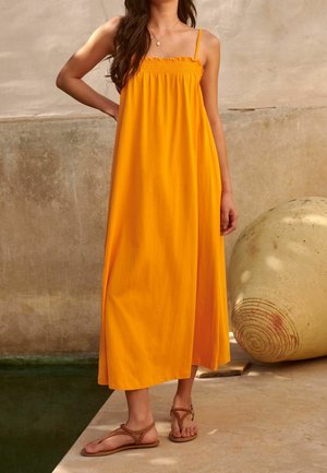 Gelbes Maxi-Kleid aus leichtem Stoff mit verstellbaren Trägern und Rüschenausschnitt. Kombiniert mit braunen Riemchensandalen.