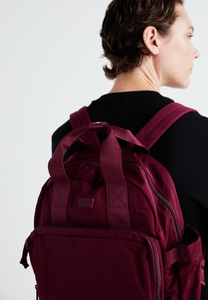 Maroon backpack with dual top handles and padded shoulder straps. Features zippered compartments and a logo patch on the front.