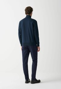 Tumši zila džemperis ar augstu apkakli, nēsājot kopā ar tumši zilām biksēm un melnām kurpēm, ar gludu tekstūru un ribotām piedurknēm.