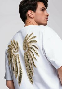 Biała koszulka z krótkim rękawem, luźnym krojem i okrągłym dekoltem, z tyłu ozdobiona złotym wzorem skrzydeł z cekinów.