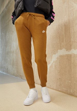 Personne portant un pantalon de survêtement Nike marron, des baskets blanches, une veste noire et un t-shirt noir avec des détails roses et bleus sur les manches, debout à l'intérieur.