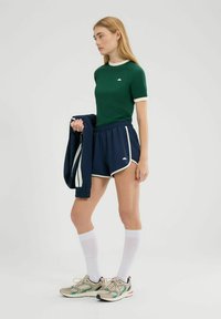Groen nauwsluitend T-shirt met witte kraag, marineblauwe shorts met witte rand, en lichte sneakers. Model houdt een marineblauwe jas vast en draagt witte sokken.