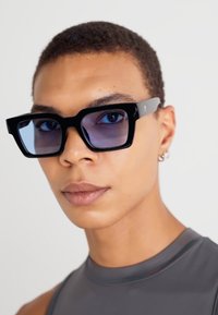 Ochelari de soare dreptunghiulari negri cu lentile colorate, având rame groase și un design elegant. Accentele includ un balama laterală de culoare gri deschis.
