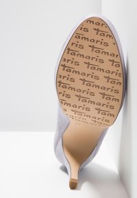 Chaussure à talons hauts en daim lavande, inclinée contre un mur, montrant la semelle avec le texte répétitif "tamaris" formant un motif antidérapant.