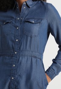 Jeansskjol med långa ärmar, knappstängning fram, två bröstfickor, samlad midja och en medelblå nyans.