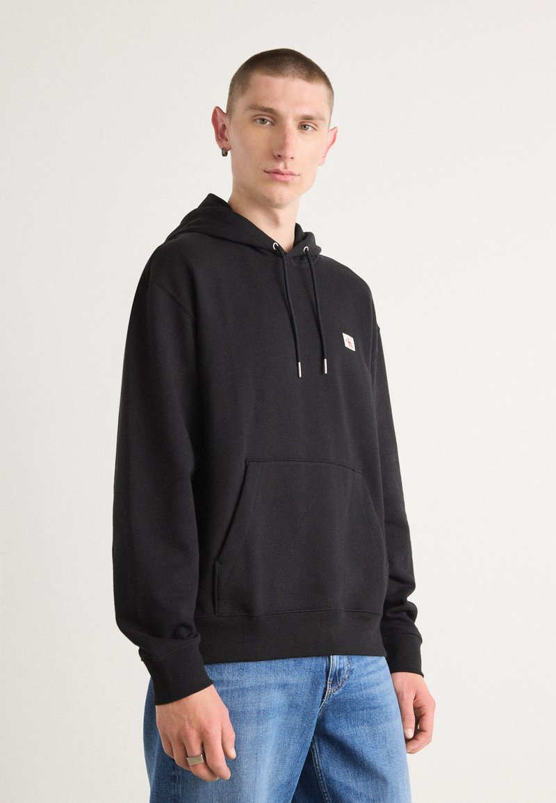 Zwarte pullover hoodie met een voorkant kangaroo-zak en trekkoorden. Beschikt over een klein logo-badge en geribbelde manchetten, gecombineerd met blauwe spijkerbroek.