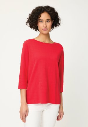 Kobieta z krótkimi kręconymi włosami, ubrana w czerwoną bluzkę z długim rękawem i białe spodnie, stojąca na tle jednolitego jasnego tła.