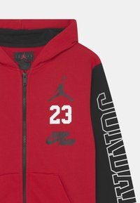 Czerwona i czarna bluza z kapturem na suwak, z kapturem, z białym nadrukiem "23" oraz logo Jumpman, a na rękawie odważny napis "JORDAN". Miękka tkanina.