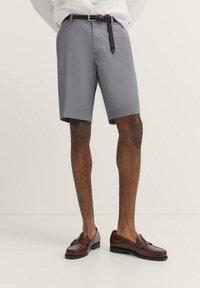 Navy Chino-Shorts mit einem Rautenmuster, ausgestattet mit einem Reißverschluss und Knopfverschluss. Kombiniert mit braunen Lederschuhen und einem geflochtenen Gürtel.