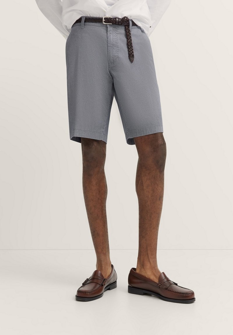 Navy Chino-Shorts mit einem Rautenmuster, ausgestattet mit einem Reißverschluss und Knopfverschluss. Kombiniert mit braunen Lederschuhen und einem geflochtenen Gürtel.