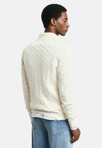 Pull en maille torsadée crème avec un col et des poignets côtelés, présentant une coupe décontractée et un motif texturé. Porté par-dessus une chemise rayée.