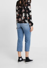 Blouse noire à motif floral avec un détail en dentelle dans le dos ; associée à un jean court bleu clair et des baskets montantes noires à semelle blanche.