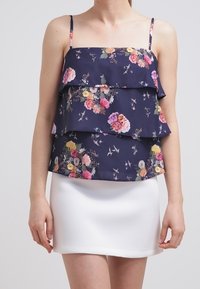Námořnický květinový top s vrstvenými volány a tenkými ramínky, zdobený růžovými, žlutými a bílými květinami na tmavém pozadí, doplněný bílou sukni.