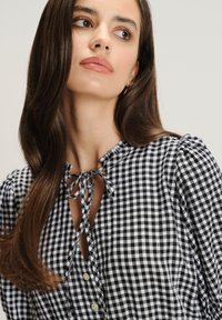 Donna con lunghi capelli castani che guarda verso destra, indossando una blusa a scacchi bianca e nera con collo annodato e bottoni.