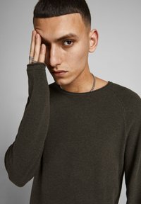 Donkergroene gebreide trui met een gestructureerde weving, ronde halslijn en lange mouwen. Model poseert met een hand gedeeltelijk voor haar gezicht.