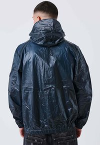 Uomo con i capelli corti che indossa una giacca a vento con cappuccio lucida di colore blu scuro, di spalle, abbinata a jeans in denim scuro.