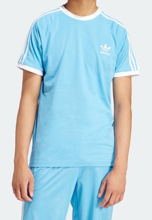 Osoba oblečená do světle modrého sportovního oblečení Adidas s bílými detaily, logem na hrudi a třemi bílými pruhy běžícími po rukávech a kalhotách.