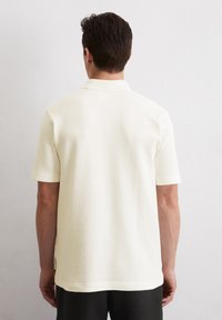 Cremfarbendes Polohemd mit strukturierter Oberfläche, ribbedem Kragen und kurzen Ärmeln. Es hat einen geraden Saum und ein seitliches Label-Detail.