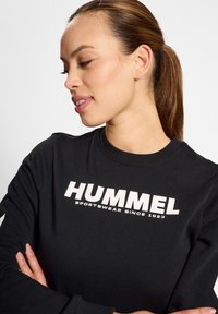 Czarny sweter z logo "HUMMEL SPORTSWEAR SINCE 1923" w kolorze białym. Widoczny ribbowany okrągły dekolt i długie rękawy.