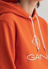 Sweat à capuche orange vif en tissu doux, avec des cordons blancs et un logo brodé. Le design comprend une poche kangourou à l'avant.
