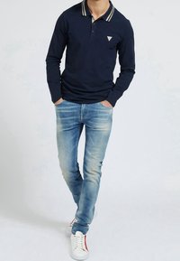 Man wearing navy long-sleeve polo shirt with stripe collar, light blue faded jeans, white sneakers, standing with hands in pockets.