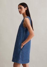 Denim shift-jurk met mouwloos ontwerp, zijzakken en rechte snit. Kleur is medium blauw met zichtbare stikdetails.