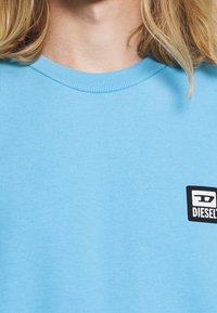 Světle modrý crew-neck sweatshirt vyrobený z měkké bavlny, s malou černou nášivkou loga Diesel na levé části hrudi.