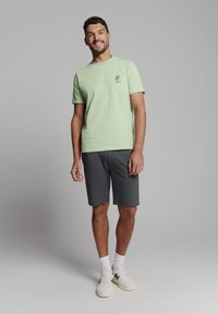 Lichtgroene t-shirt met korte mouwen en een kleine grafische print op de borst, gecombineerd met donkergrijze sportshorts en witte sneakers.
