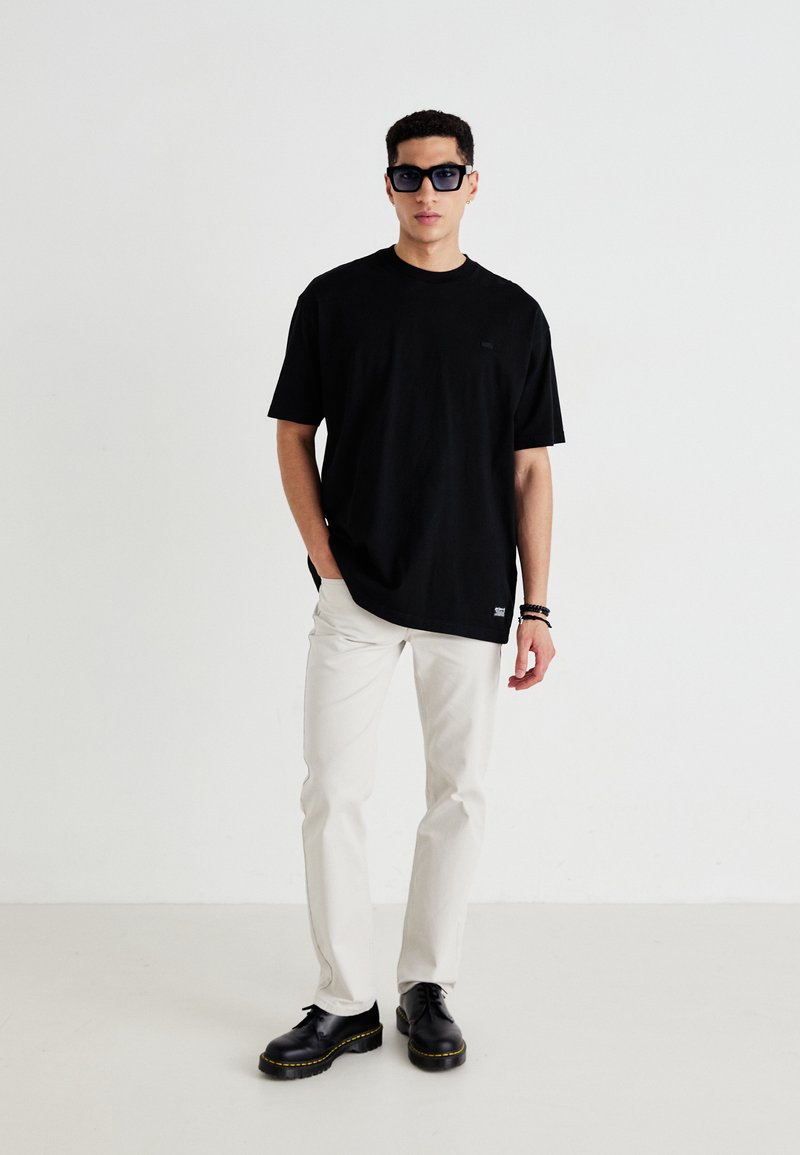 Czarna bawełniana koszulka z krótkim rękawem, beżowe spodnie i czarne sznurowane buty. Dodatki to ciemne okulary przeciwsłoneczne. Minimalistyczny design.