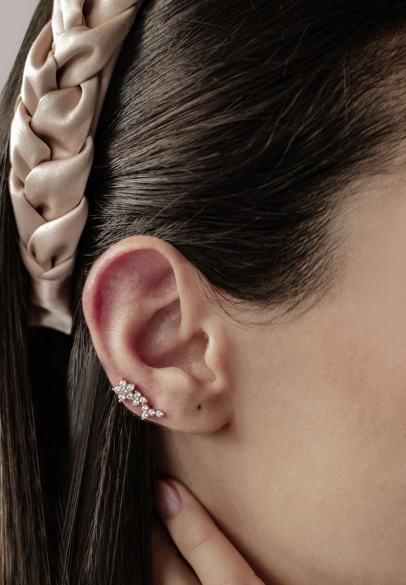 Srebrny klips do ucha z wieloma małymi przezroczystymi kamieniami ułożonymi w kwiatowy wzór, przymocowany do płatka ucha, z ciemnymi włosami w tle.