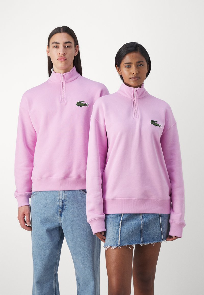 Felpe in pile rosa con colli alti, dotate di un logo ricamato verde. Indossate con jeans in denim blu e una gonna in denim str appata.