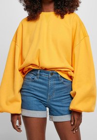 Sudadera amarilla de gran tamaño con mangas caídas, combinada con pantalones cortos de mezclilla de talle alto con dobladillo vuelto. Textura suave, ajuste relajado.