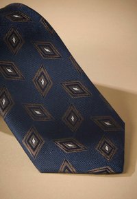Navy tie with a textured surface featuring overlapping diamond patterns in brown and silver, displayed against a beige background.