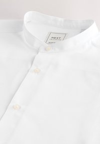 Bijela košulja s ovratnikom od glatkog tkanja, s prednjim dijelom s kopčanjem na gumbe od bisera i etiketom s natpisom "NEXT TAILORING."