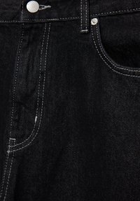 Gros plan sur un jean en denim noir montrant la poche avant, un rivet argenté, un bouton et des détails de couture blanche.